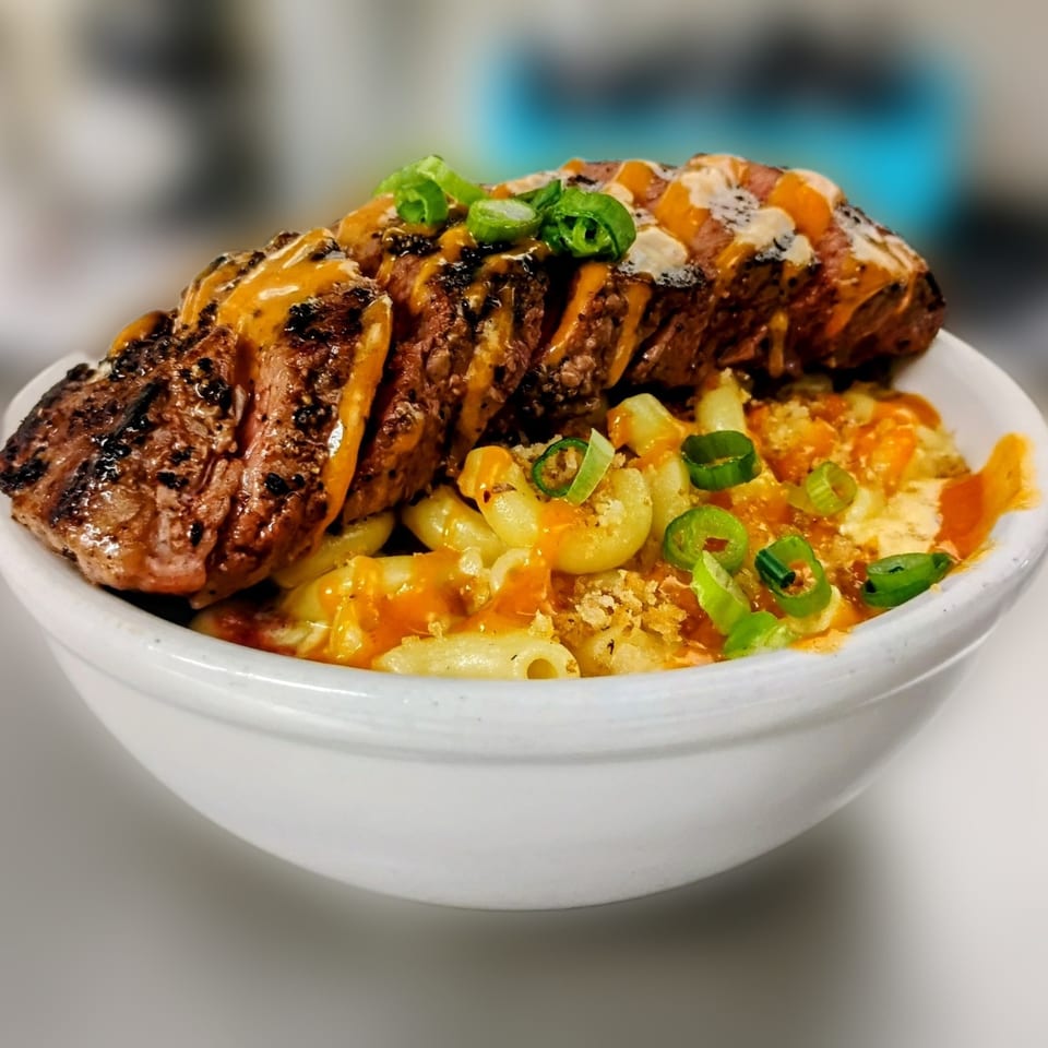 Grilled steak slices with sauce and green onions over macaroni and cheese in a white bowl.