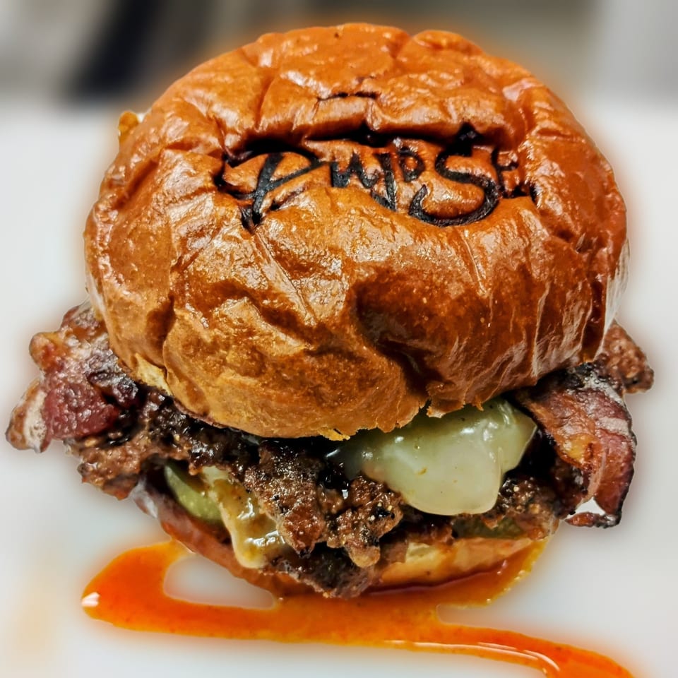 Pujo St Cafe burger with a branded bun, bacon, cheese, and sauce on a white plate.